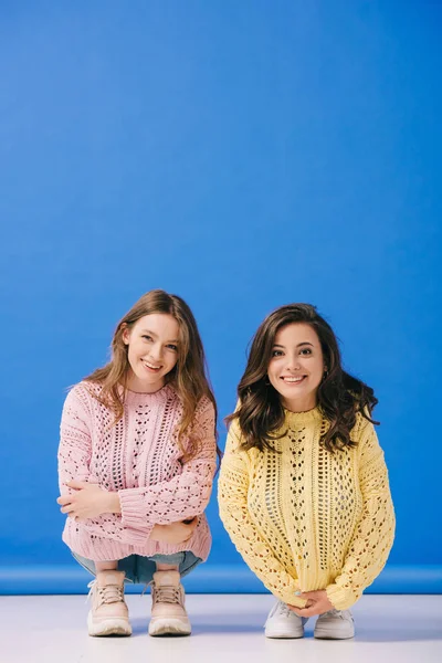 Mujeres atractivas y sonrientes en suéteres mirando a la cámara sobre fondo azul - foto de stock