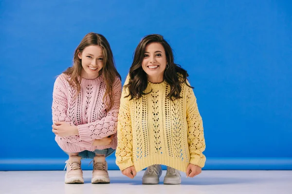 Mujeres atractivas y sonrientes en suéteres mirando a la cámara sobre fondo azul - foto de stock