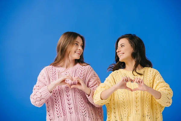 Attractive and smiling women in sweaters showing heart gesture isolated on blue — Stock Photo