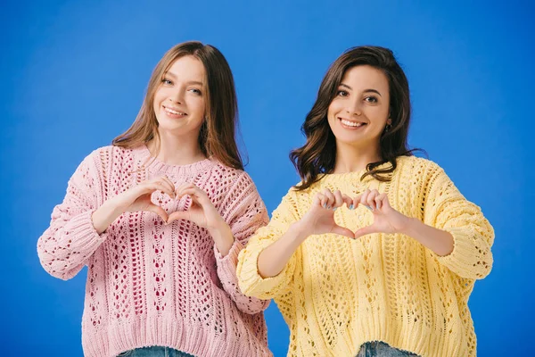 Привлекательные и улыбающиеся женщины в свитерах, показывающие жест сердца, изолированный на голубом — стоковое фото