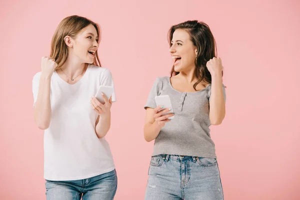 Mujeres atractivas y sonrientes en camisetas con teléfonos inteligentes y mostrando sí gesto aislado en rosa - foto de stock