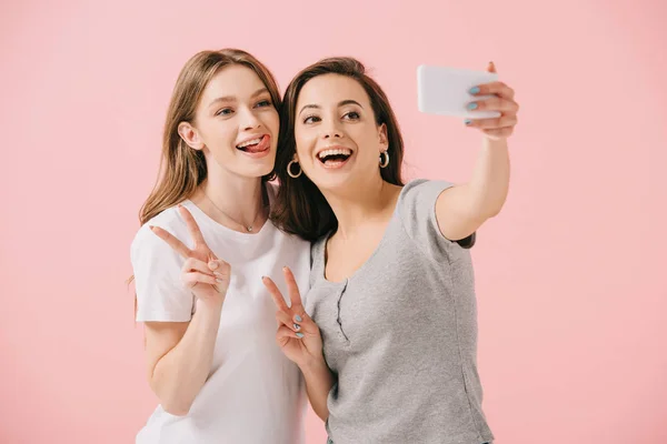 Mujeres atractivas y sonrientes en camisetas tomando selfie y mostrando signos de paz aislados en rosa - foto de stock