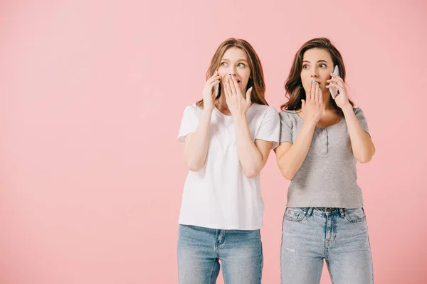 Привабливі і шоковані жінки в футболках розмовляють на смартфонах ізольовані на рожевому — стокове фото