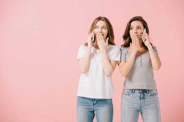 Mujeres atractivas y conmocionadas en camisetas hablando en teléfonos inteligentes aislados en rosa - foto de stock