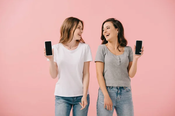 Lächelnde und attraktive Frauen in T-Shirts, die Smartphones isoliert auf rosa halten — Stockfoto