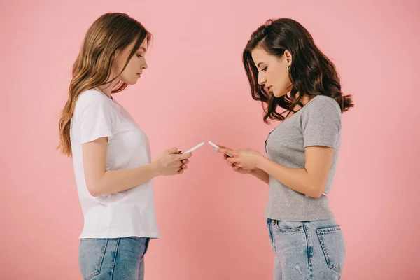 Vista lateral de mujeres atractivas en camisetas usando teléfonos inteligentes sobre fondo rosa - foto de stock