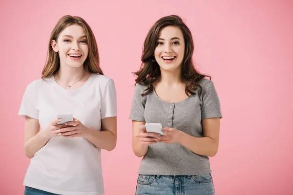 Привабливі і усміхнені жінки в футболках тримають смартфон ізольовані на рожевому — стокове фото