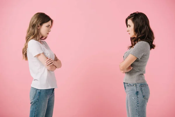 Attractive and serious women in t-shirts with crossed arms looking at each other isolated on pink — Stock Photo