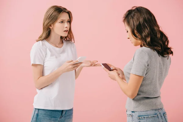 Mulher atraente e chocado em t-shirt segurando smartphone e olhando para seu amigo isolado em rosa — Fotografia de Stock