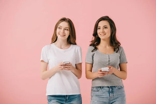 Привлекательные и улыбающиеся женщины в футболках, держащие смартфоны изолированными на розовом — стоковое фото