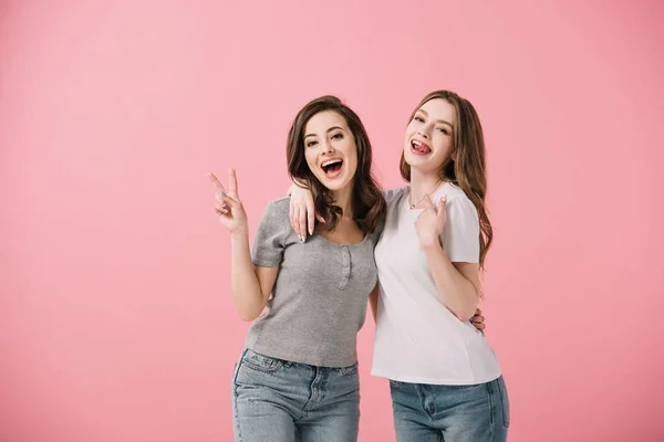 Привлекательные и улыбающиеся женщины в футболках, смотрящие в камеру и показывающие знак мира, изолированный на розовом — стоковое фото