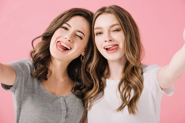 Attraktive und lächelnde Frauen in T-Shirts mit herausgestreckten Zungen, die vereinzelt in die Kamera auf rosa sehen — Stockfoto