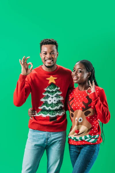 Africano americano homem e mulher em camisolas de Natal mostrando sinal ok e olhando para a câmera isolada no verde — Fotografia de Stock