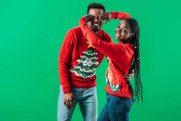 Mulher afro-americana em camisola de Natal cobrindo os olhos com as mãos para o homem isolado no verde — Fotografia de Stock