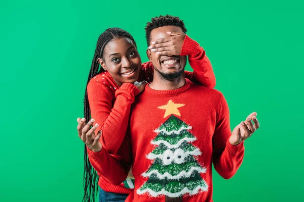Mulher afro-americana em camisola de Natal cobrindo os olhos com a mão para o homem isolado no verde — Fotografia de Stock