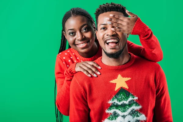 Femme afro-américaine en pull de Noël tenant la main près des yeux de l'homme isolé sur vert — Photo de stock