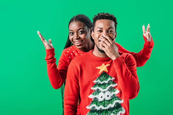 African American woman raising hands behind surprised man in Christmas sweater isolated on green — Stock Photo