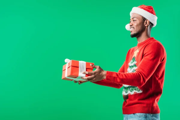 Side view of African American man in Christmas sweater and Santa hat holding red gift box looking away isolated on green — Stock Photo