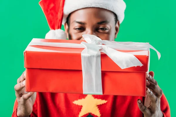 African American man in Santa hat holding red gift box with white ribbon isolated on green — Stock Photo