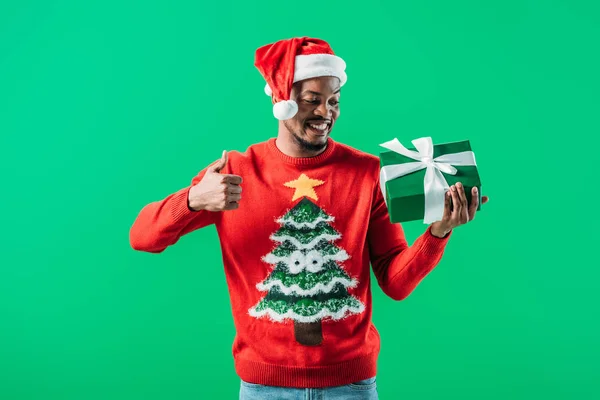 African American man in Christmas sweater and Santa hat showing thumb up and looking at gift box in his hands isolated on green — Stock Photo