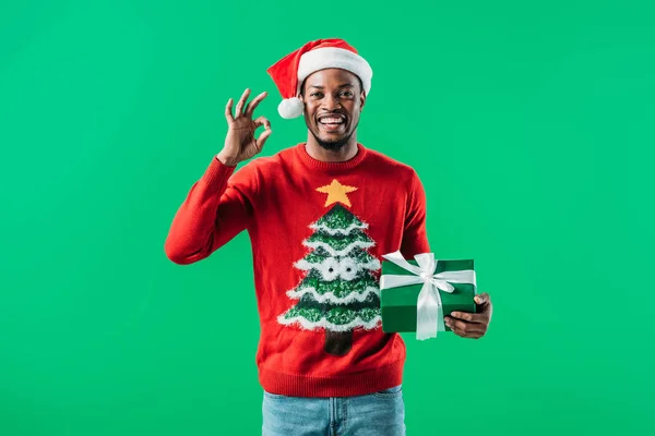 Afrikanisch-amerikanischer Mann in Weihnachtspullover und Weihnachtsmütze zeigt Okay Zeichen und hält Geschenkbox isoliert auf grün — Stockfoto