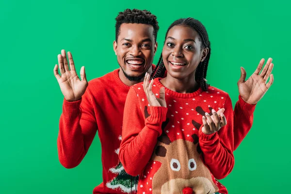 Счастливая афроамериканская пара в красных рождественских свитерах улыбается и смотрит на камеру, изолированную на зеленый — стоковое фото