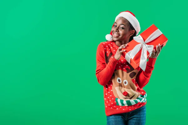 Афроамериканка в рождественском свитере и шляпе Санты держит подарочную коробку и смотрит на камеру, изолированную на зеленый — стоковое фото