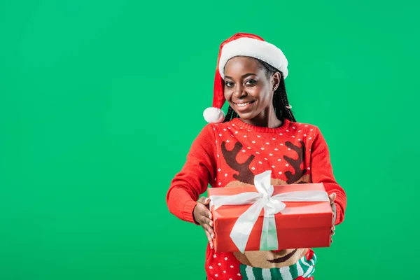 African American woman in Christmas sweater and Santa hat holding gift box with white ribbon and looking at camera isolated on green — Stock Photo