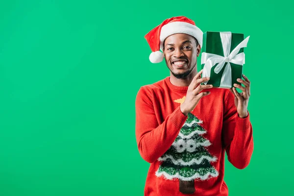 Afrikanisch-amerikanischer Mann in Weihnachtspullover und Weihnachtsmütze mit Geschenk und Blick in die Kamera isoliert auf grün — Stockfoto