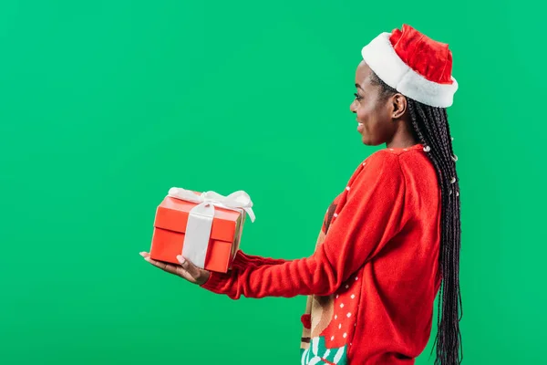Vista lateral de la mujer afroamericana en suéter de Navidad y sombrero de Santa levantando las manos con caja de regalo aislado en verde - foto de stock