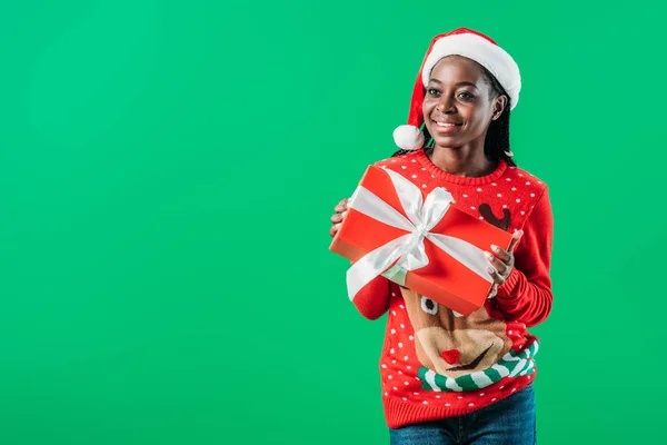 African American woman in Christmas sweater and Santa hat holding present isolated on green — Stock Photo