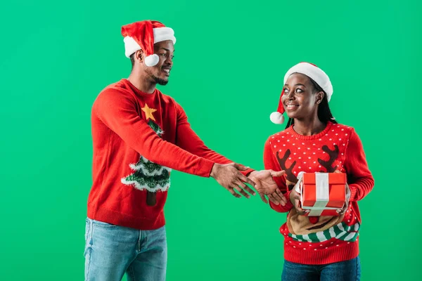 Hombre afroamericano en jersey rojo de Navidad levantando las manos a la caja de regalo en manos de mujer aislada en verde - foto de stock
