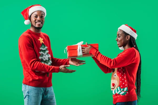 Side view of African American woman in red Christmas sweater giving gift box to man isolated on green — Stock Photo