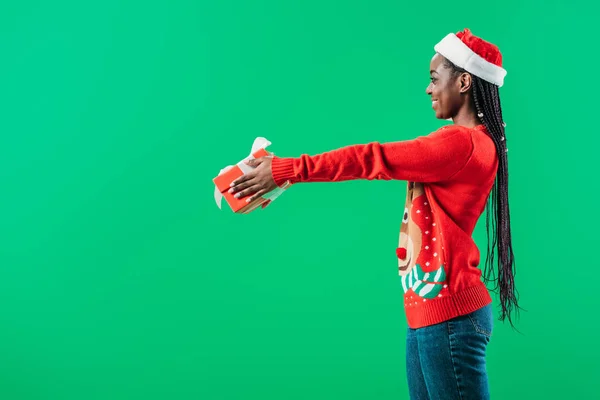 Side view of African American woman in Christmas sweater and Santa hat raising hands with gift box isolated on green — Stock Photo
