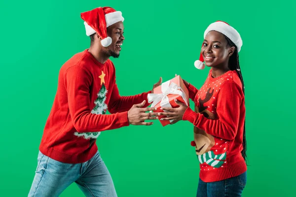 Side view of African American man in red Christmas sweater and Santa hat getting gift box from woman isolated on green — Stock Photo