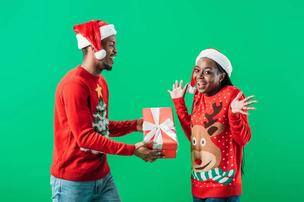 Афроамериканець людина в червоному Різдво светр і Санта-Hat даючи Подарункова коробка здивована жінка, ізольована на зелений — стокове фото