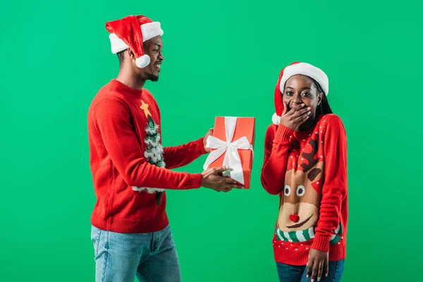 African American man in red Christmas sweater giving gift box to surprised woman who covering mouth with hand isolated on green — Stock Photo