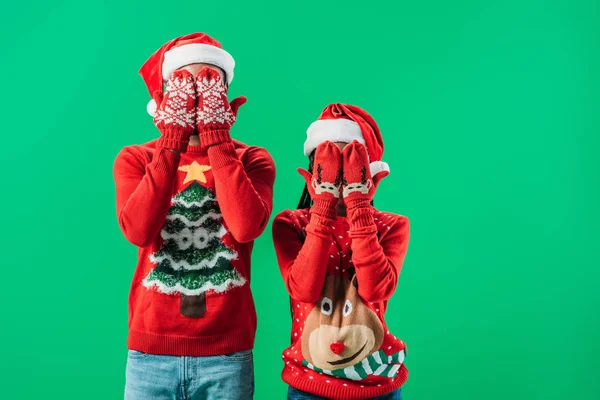 Casal afro-americano em camisolas vermelhas de Natal e chapéus de Papai Noel cobrindo os olhos com as mãos em luvas de inverno isolado em verde — Fotografia de Stock