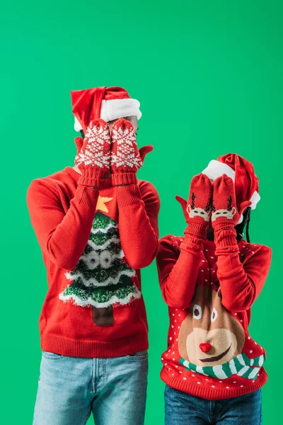 Афро-американская пара в красных рождественских свитерах и шляпах Санты, закрывающих глаза руками в зимних перчатках с узором, изолированным на зеленом — стоковое фото