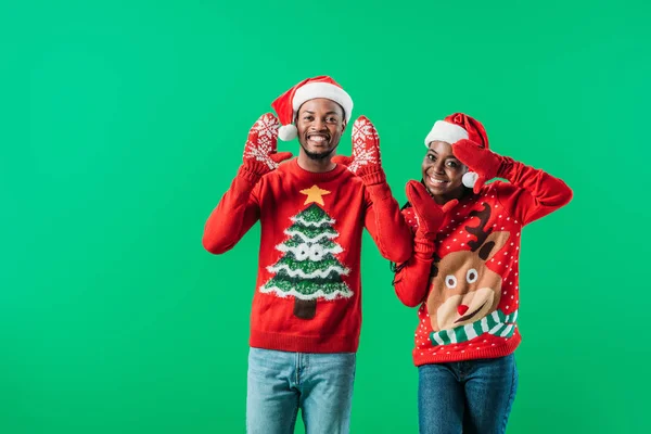 African American couple in red Christmas sweaters and Santa hats raising hands in winter gloves with pattern isolated on green — Stock Photo