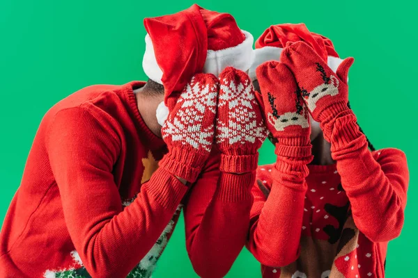 Couple afro-américain en pulls de Noël et chapeaux de Père Noël couvrant les yeux avec les mains en gants rouges d'hiver avec motif isolé sur vert — Photo de stock