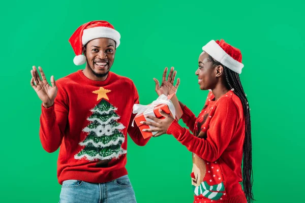 Afrikanisch-amerikanische Frau in rotem Weihnachtspullover und Weihnachtsmütze schenkt Geschenkbox an überraschten Mann isoliert auf grün — Stockfoto