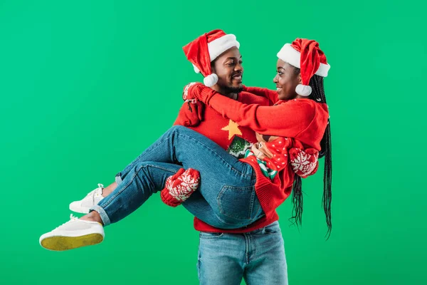 Afrikanisch-amerikanischer Mann in rotem Weihnachtspullover und Weihnachtsmütze hält lächelnde Frau isoliert auf grünem Grund — Stockfoto