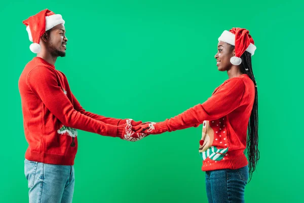 Seitenansicht eines afrikanisch-amerikanischen Paares in roten Weihnachtspullovern und Weihnachtsmützen, die sich die Hände in Handschuhen halten und sich isoliert auf grün betrachten — Stockfoto