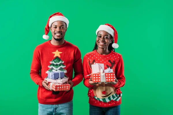 Afrikanisches amerikanisches Paar in roten Weihnachtspullovern und Weihnachtsmützen mit Geschenken und Blick in die Kamera isoliert auf grün — Stockfoto