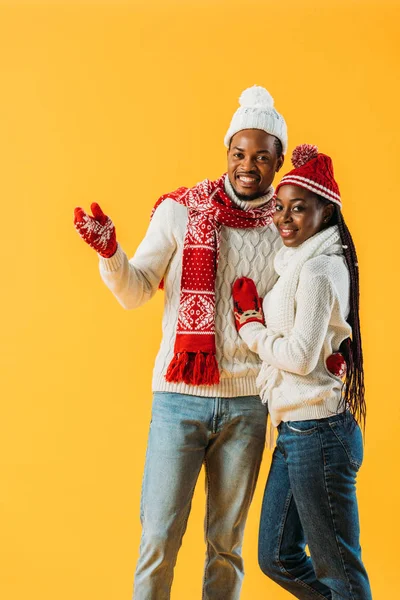 African American man in cozy winter outfit hugging woman and pointing with hand isolated on yellow — Stock Photo