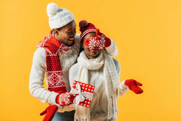 Homem afro-americano em roupas de inverno acolhedor segurando caixa de presente e cobrindo com olhos de mão de mulher isolada no amarelo — Fotografia de Stock