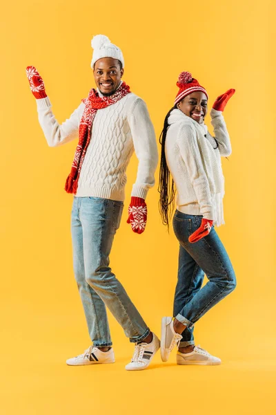 African American couple in winter outfit standing back to back, smiling and looking at camera isolated on yellow — Stock Photo