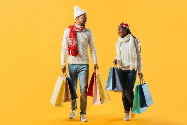 African American couple in winter outfit walking with shopping bags and looking at each other isolated on yellow — Stock Photo