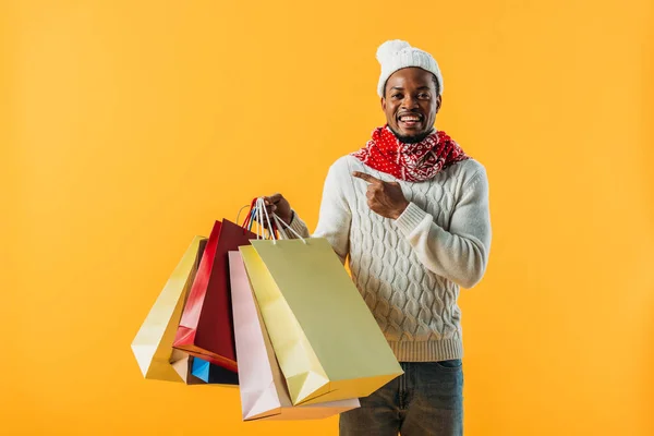 Hombre afroamericano en traje de invierno sosteniendo bolsas de compras y señalando con el dedo aislado en amarillo - foto de stock